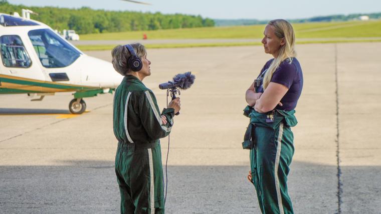 Photo of instructor Elaine Grant outside on the tarmac at an airport. She's wearing headphones and holding a digital recorder with a fuzzy covering up as she interviews flight nurse Denise Saucier. Both wear dark green flight suits with one white stripe down the leg. The sky is blue and clear. Behind the two women is the nose of a white helicopter. 