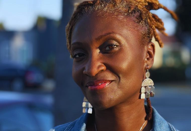 A woman wearing large silver earrings, with golden locks tied up. Wearing a denim jacket and smiling to the camera.